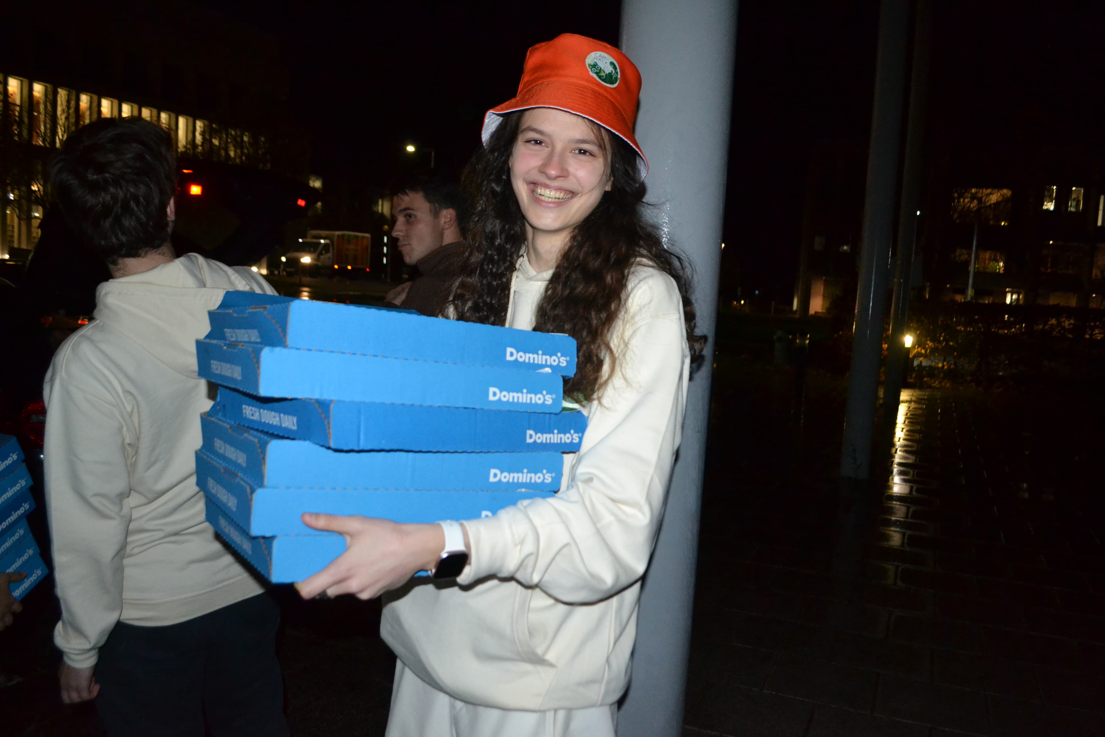 A smiling organiser in an orange bucket hat carrying a stack of Domino's pizza boxes at night
